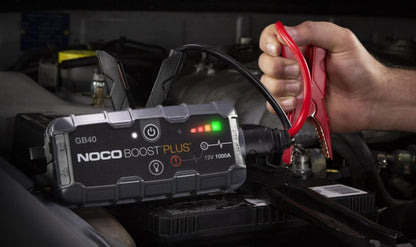 Noco Boost Plus GB40 starthjælp, rød kabelklemme til bilbatteri under motorhjelm, hånd holder klemmen.