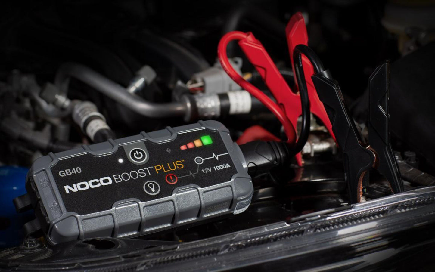 Noco Boost Plus GB40 starthjælp til bil med røde og sorte kabler på bilbatteri, grønne indikatorlys lyser.