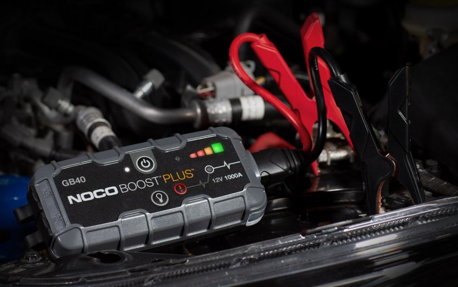 Noco Boost Plus GB40 starthjælp til bil med røde og sorte kabler på bilbatteri, grønne indikatorlys lyser.