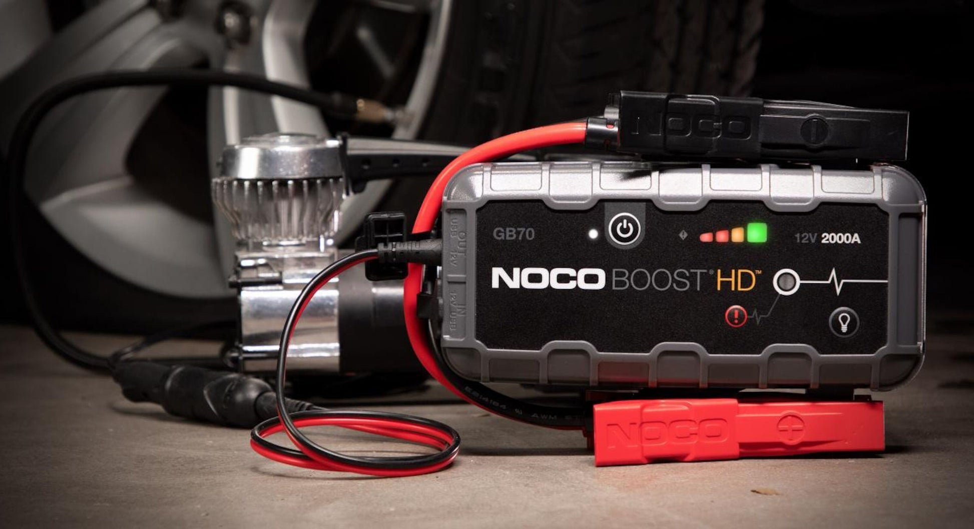 Noco Boost Plus GB70 bærbar 12V jump starter 2000A kapacitet ved siden af kompressor og bildæk i baggrunden.