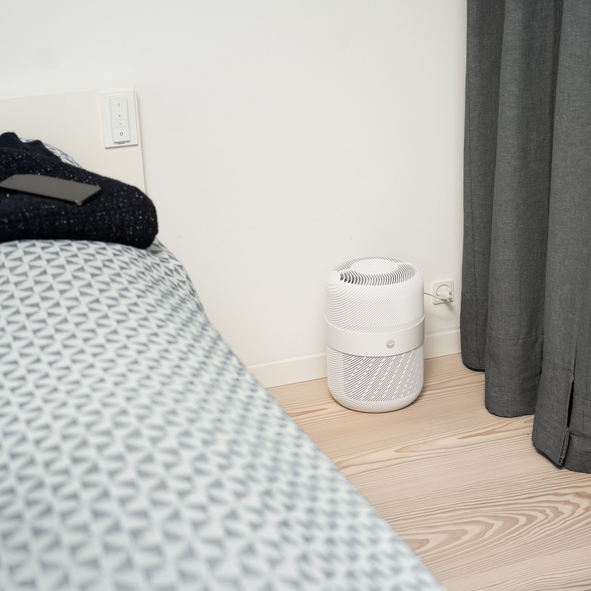 Cleverio luftrensere med HEPA-filter på lyst trægulv ved seng med mønstret dyne og grå gardin, sort telefon på seng.