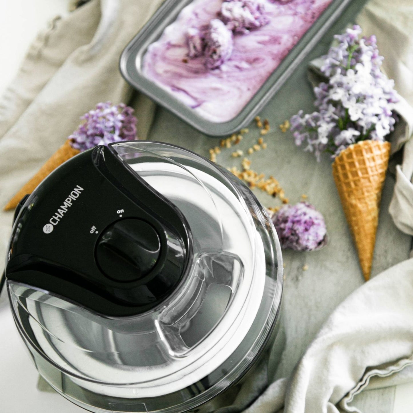 Champion Ismaskine 1,5 L sort på grå overflade med lilla frozen yoghurt i metalform og vafler pyntet med lilla blomster.