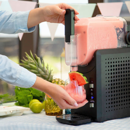 Slush ice maskine 1,9 L dispenserer lyserød slushice i glas med vandmelon pynt, frisk frugt og grønne blade på bordet.