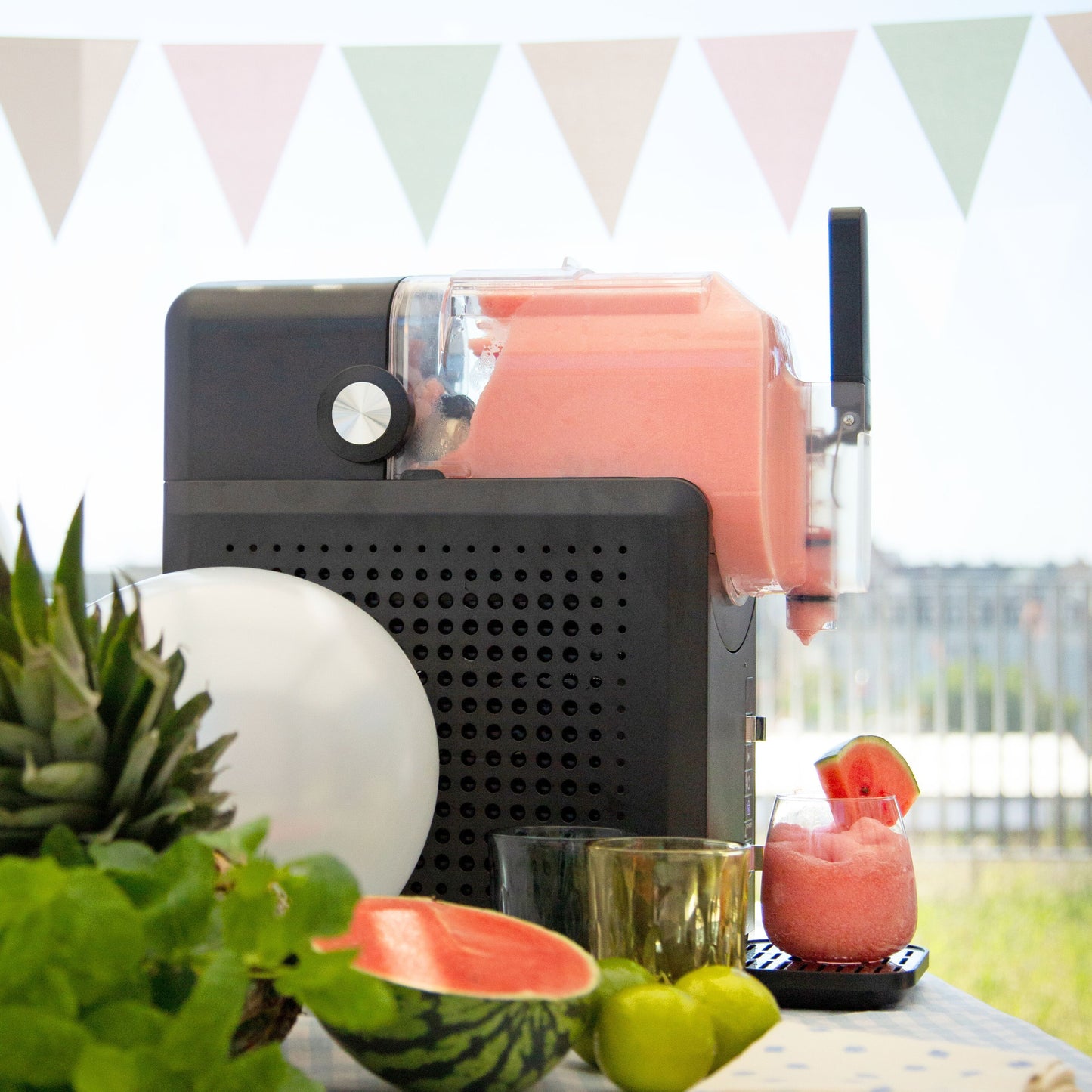 Slush ice maskine 1,9 L sort/sølv med pink slush, på bord sammen med vandmelon, ananas, lime og glas.