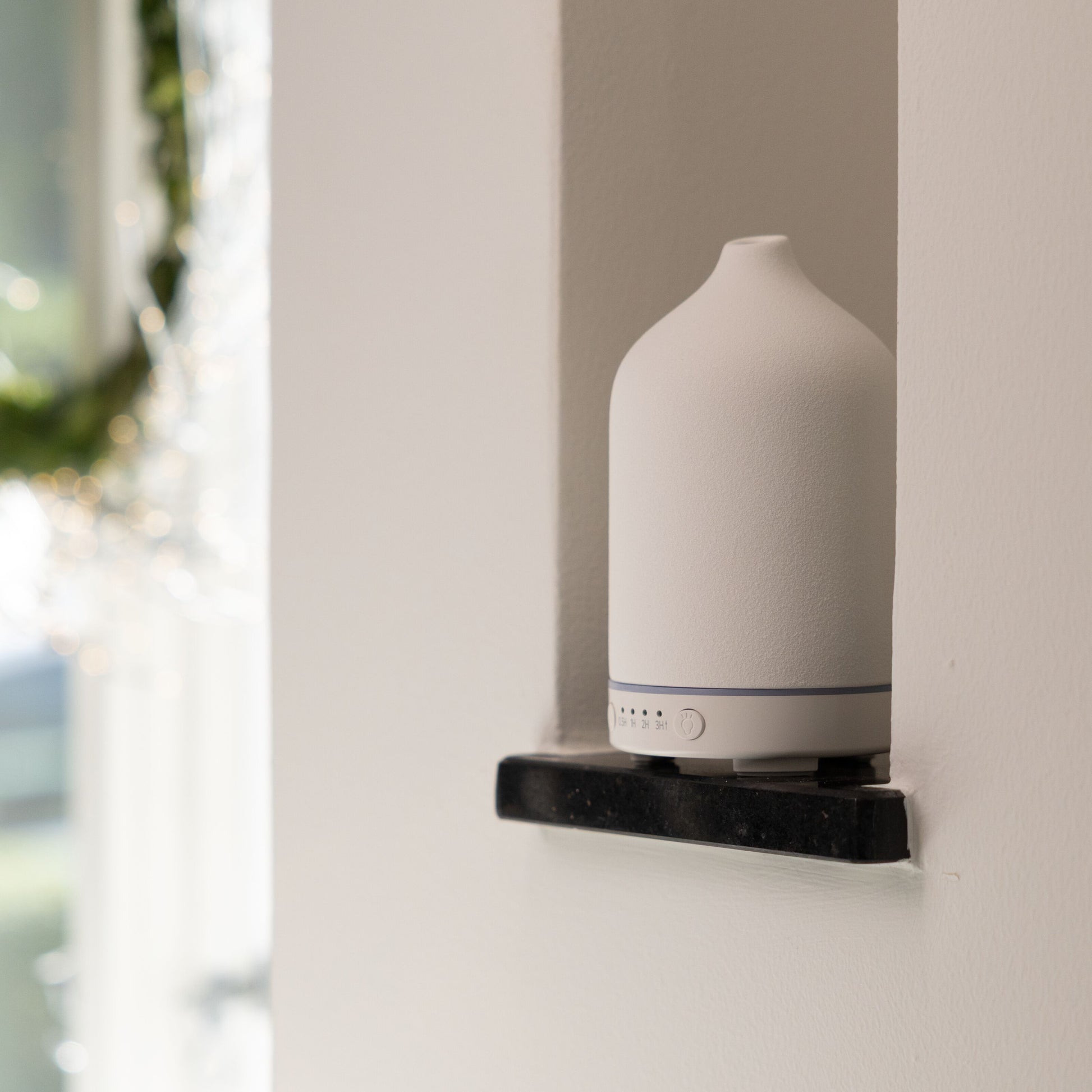 Ultralyd aromadiffuser på sort hylde i vægniche oplyst af sollys.