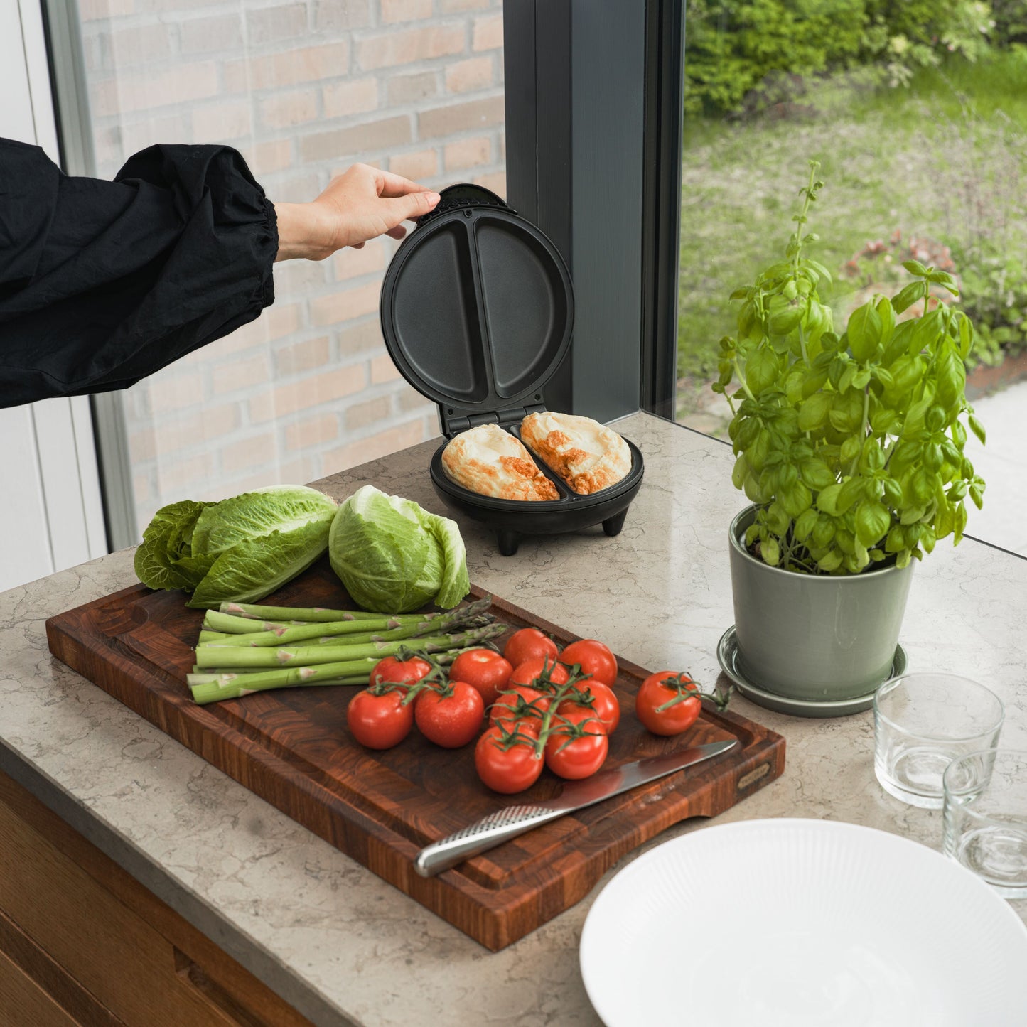 Omeletjern med non-stick belægning åbnes, to omeletter i jernet, friske tomater og asparges på skærebræt ved siden af.
