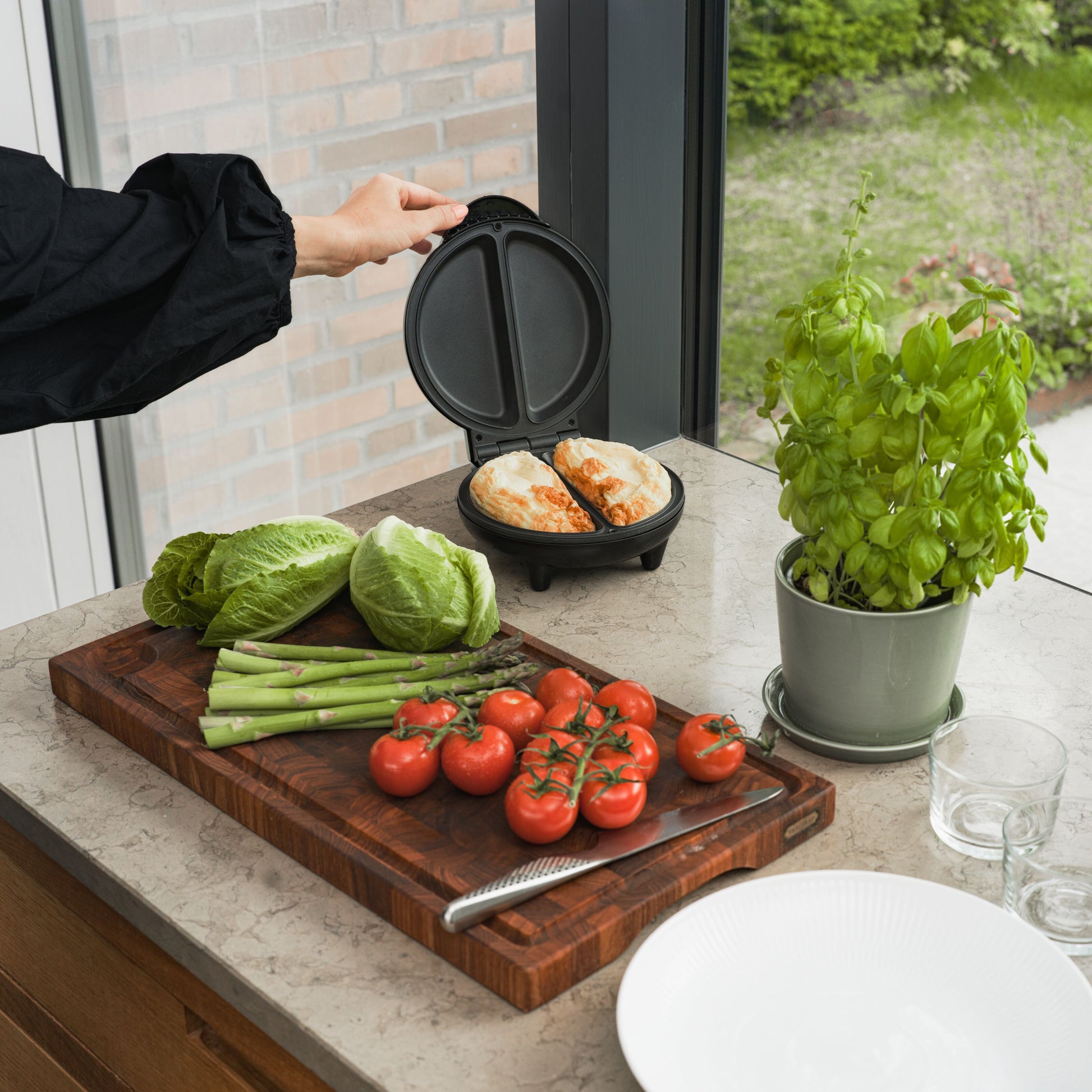 Omeletjern med non-stick belægning åbnes, to omeletter i jernet, friske tomater og asparges på skærebræt ved siden af.
