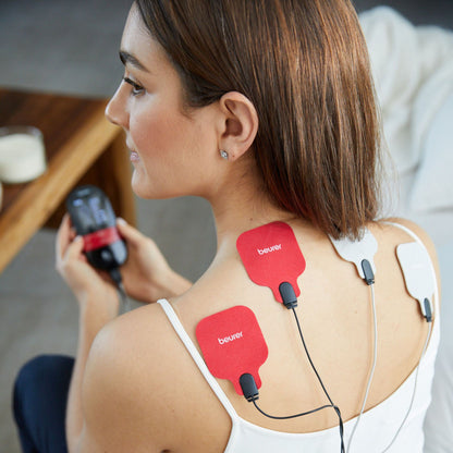 Beurer EM 59 TENS/EMS muskelstimulator med varme, røde og hvide elektrodepuder på ryg/skulder, controller i hånd.