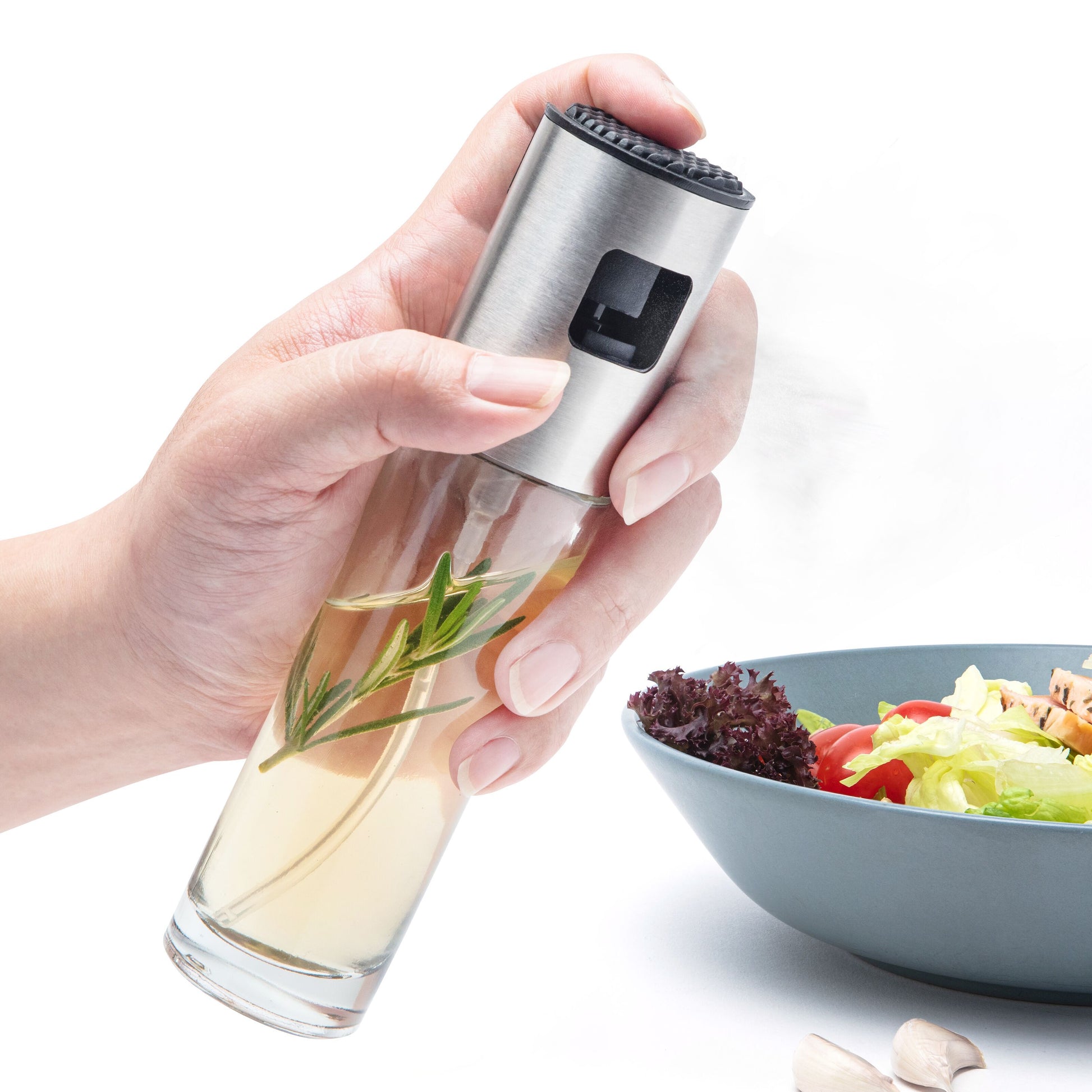 Rubicson Sprayflaske til olie med krydderurter dugger over salat i blå skål, hvidløgsfed ved siden af.