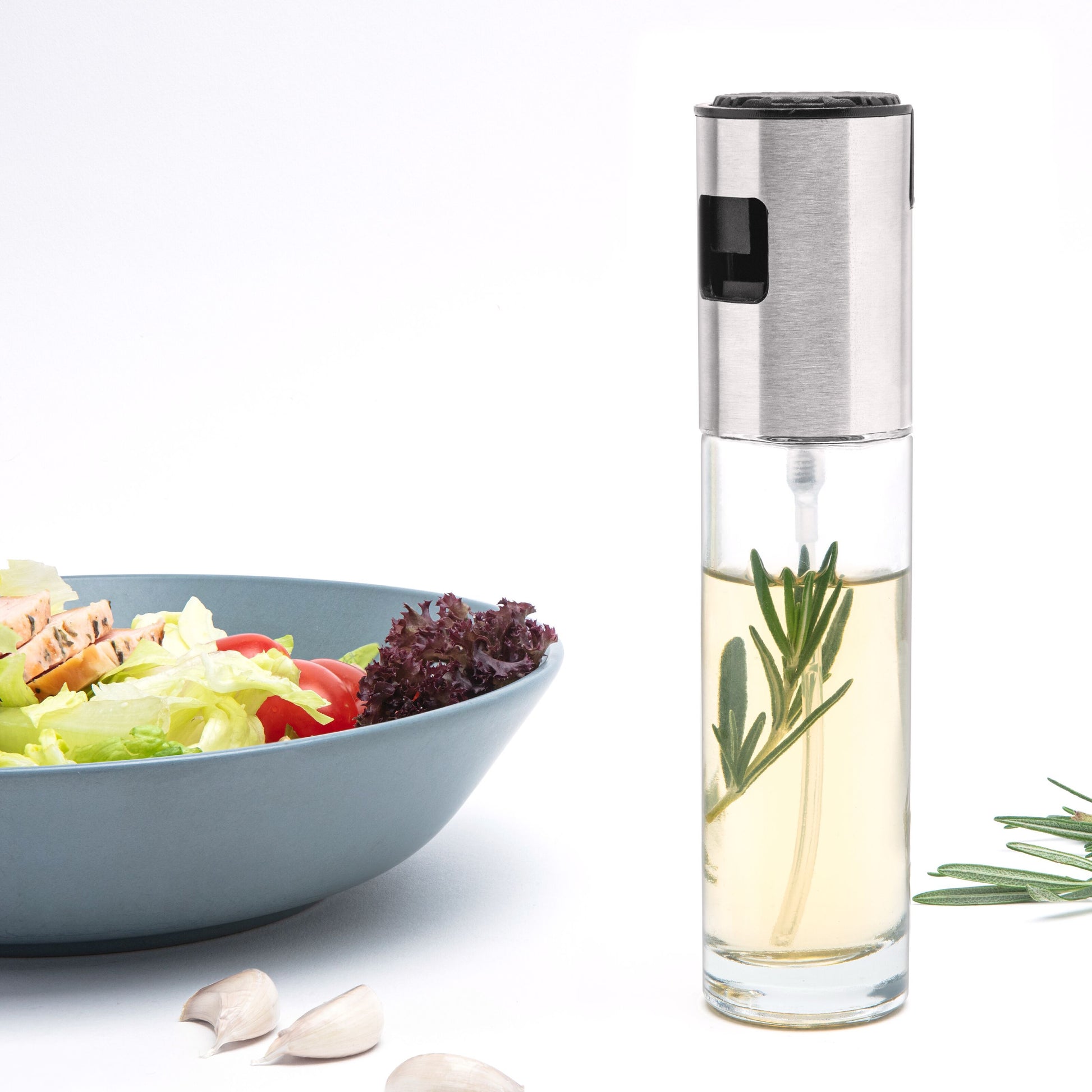 Rubicson sprayflaske til olie ved siden af blå skål med salatblade, tomat, kylling, hvidløgsfed og rosmarin.