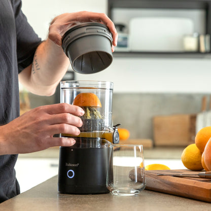 Rubicson Bærbar citruspress presser appelsin i transparent beholder, glasjuice og flere appelsiner synlige på køkkenbord.