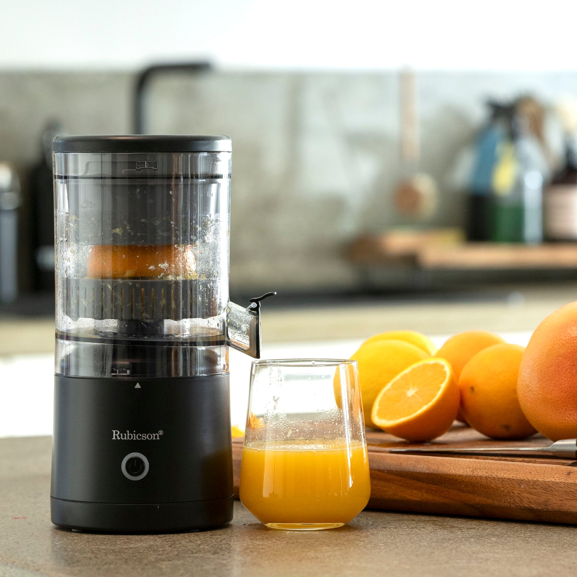 Rubicson Bærbar citruspresser sort med klar beholder ved siden af friskpresset juice og flere hele/halve appelsiner på træbræt.
