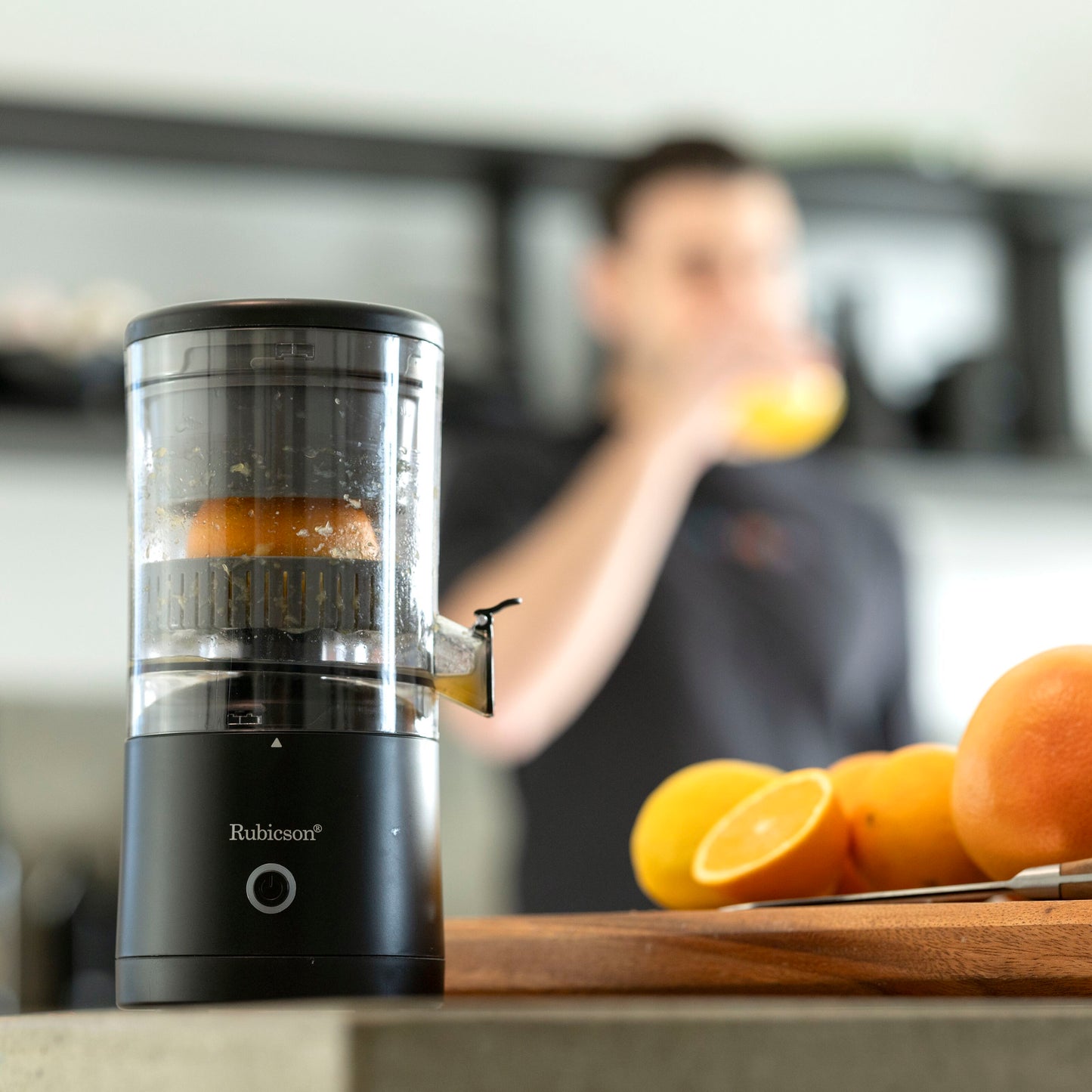 Rubicson Bærbar citruspresser presser appelsin, skiver på skærebræt, person drikker juice i baggrunden.