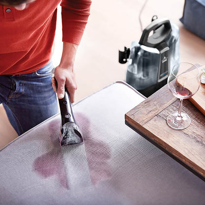 Bissel SpotClean Plus tæppe- og tekstilplejemaskine fjerner rødvinsplet på lys sofa, vin og serviet på træbord ved siden af.