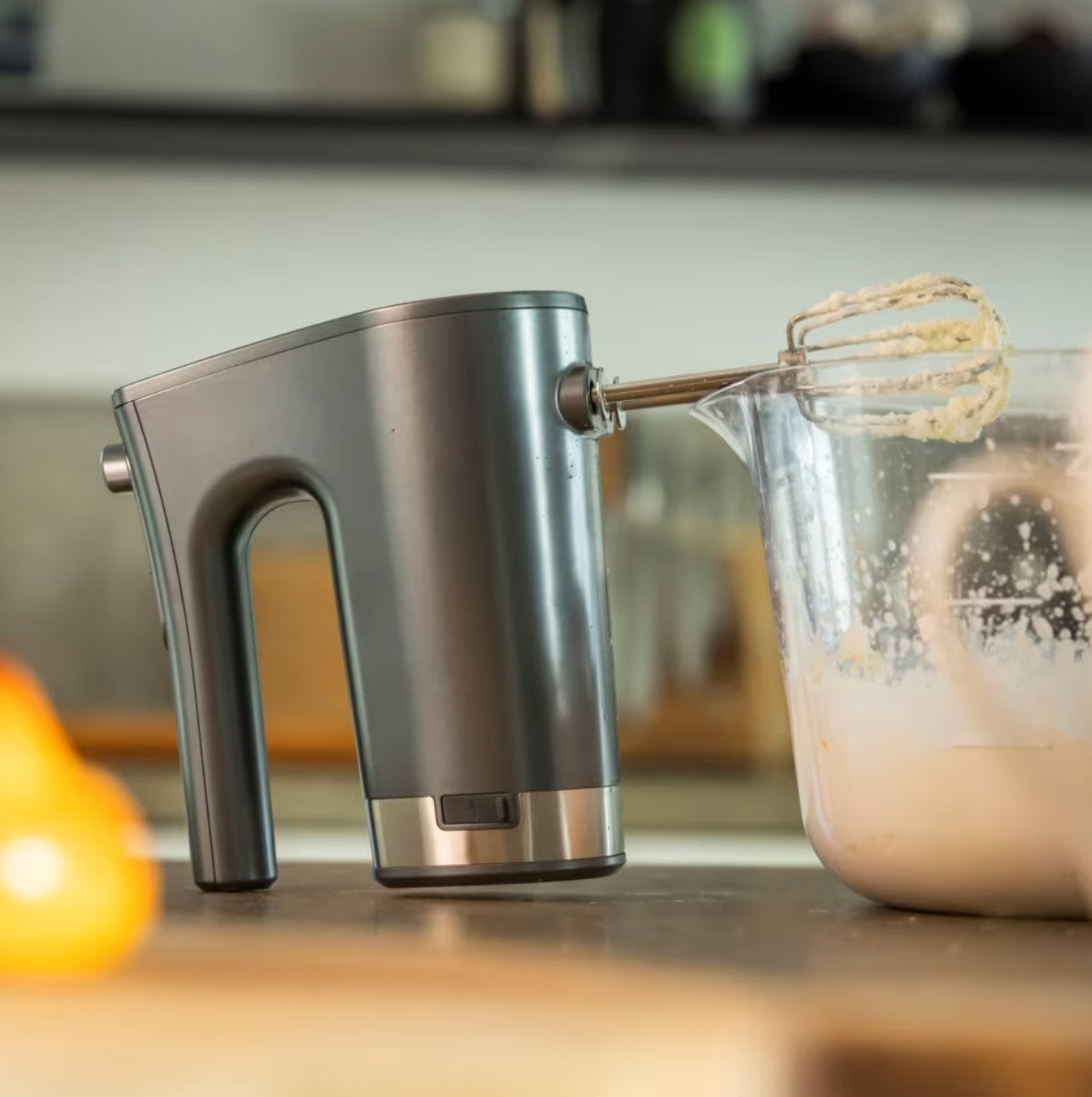 Trådløs håndmixer med piskeris ligger på kanten af klar røreskål med dej på køkkenbord.