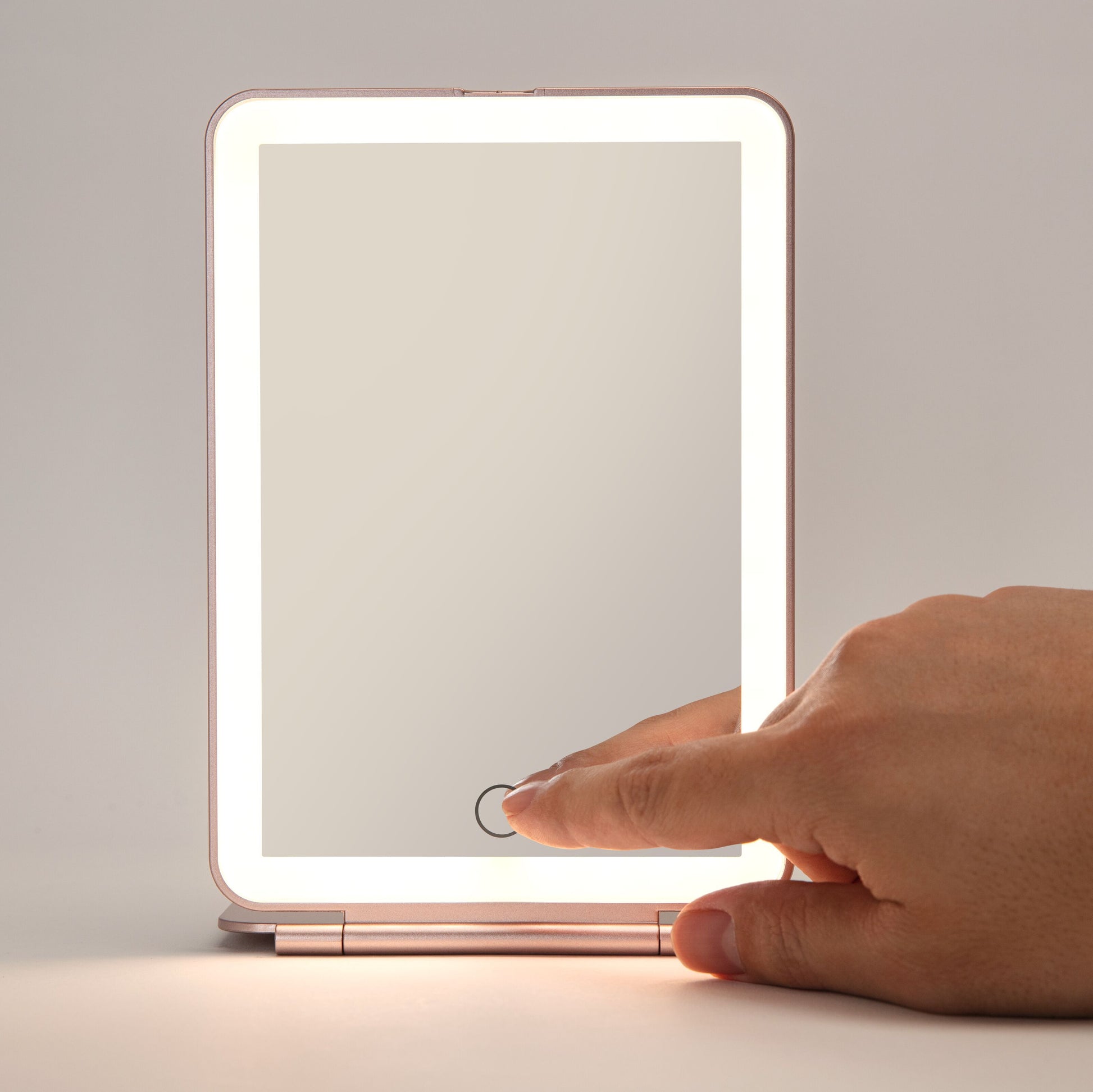 Makeup spejl med LED-belysning, rektangulært, power-knap på bunden aktiveres af hånd, står på flad overflade.