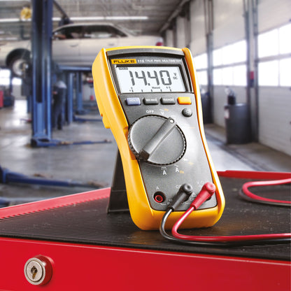 Fluke 115 multimeter 14,40 volt visning på rød overflade i autoværksted, bil løftet i baggrunden.