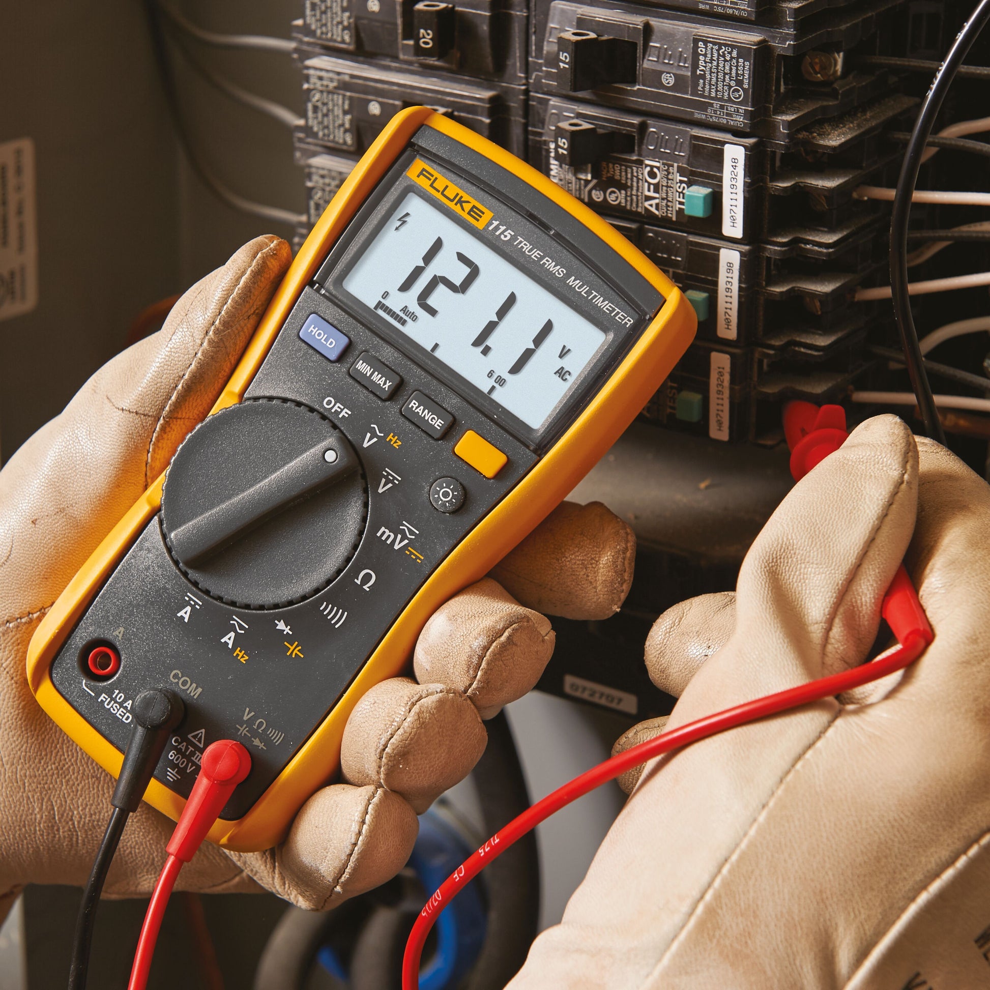 Fluke 115 multimeter viser 121,1V AC ved el-tavle med flere automatsikringer og ledninger, bruges af elektriker med håndske.