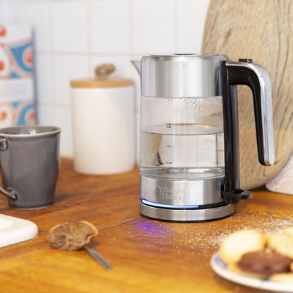 Russell Hobbs Compact Home kedel i stål på bordplade ved gråt krus, hvid beholder og tallerken med småkager.