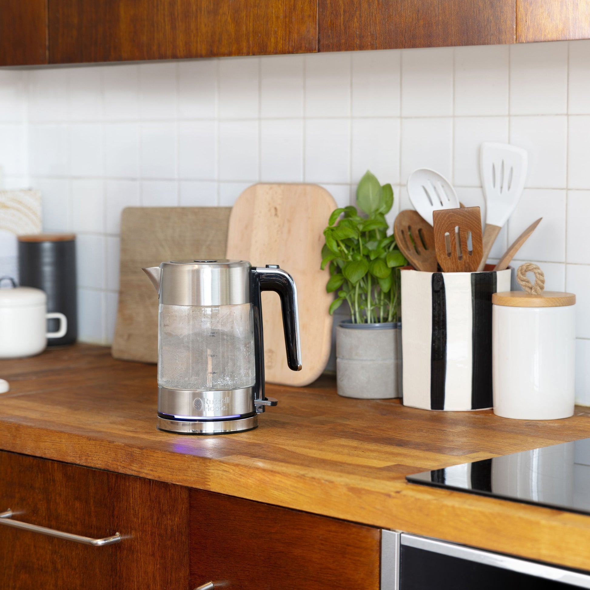 Russell Hobbs Kedel Compact Home på træbord med varmt vand, skærebrætter og køkkenredskaber foran hvidt flisebaggrund.