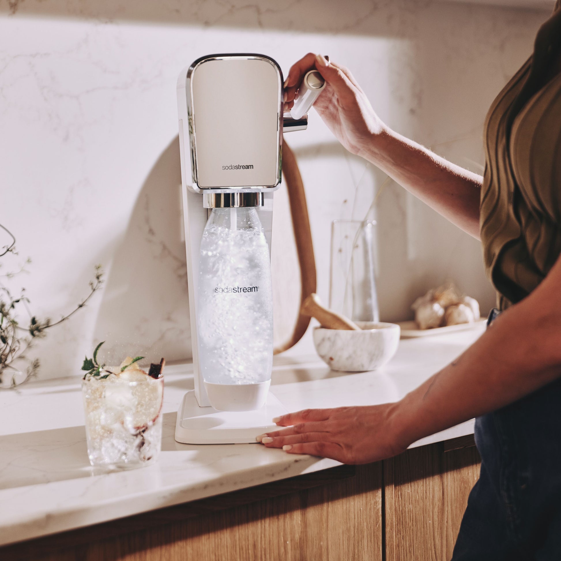 Sodastream sodavandsmaskine på køkkenbord, bruges til at lave danskvand, glas med is og garniture ved siden af.