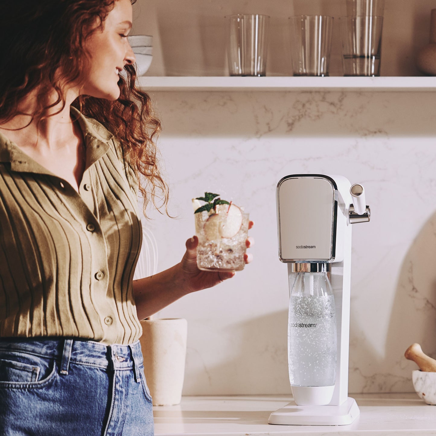 Kvinde med krøllet hår holder glas danskvand med citron og mynte ved Sodastream sodavandsmaskine på køkkenbord.