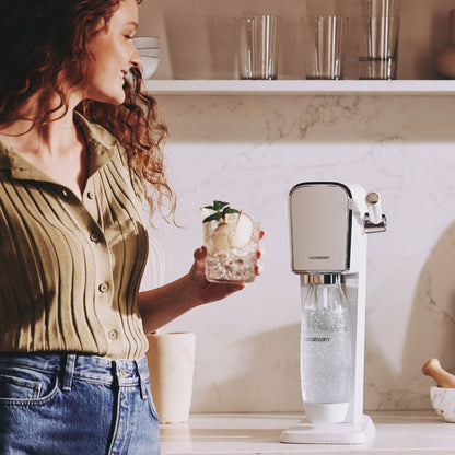 Kvinde med krøllet hår holder glas danskvand med citron og mynte ved Sodastream sodavandsmaskine på køkkenbord.