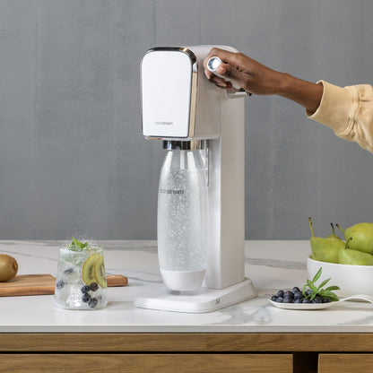 Sodastream sodavandsmaskine, sølv, kulsyreflaske monteret, frugt og danskvand synligt på bord.