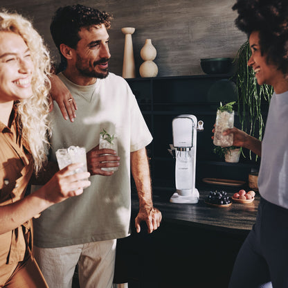 Sodastream Sodavandsmaskine på køkkenbord med friske ingredienser ses mens personer holder glas med is og mynte.