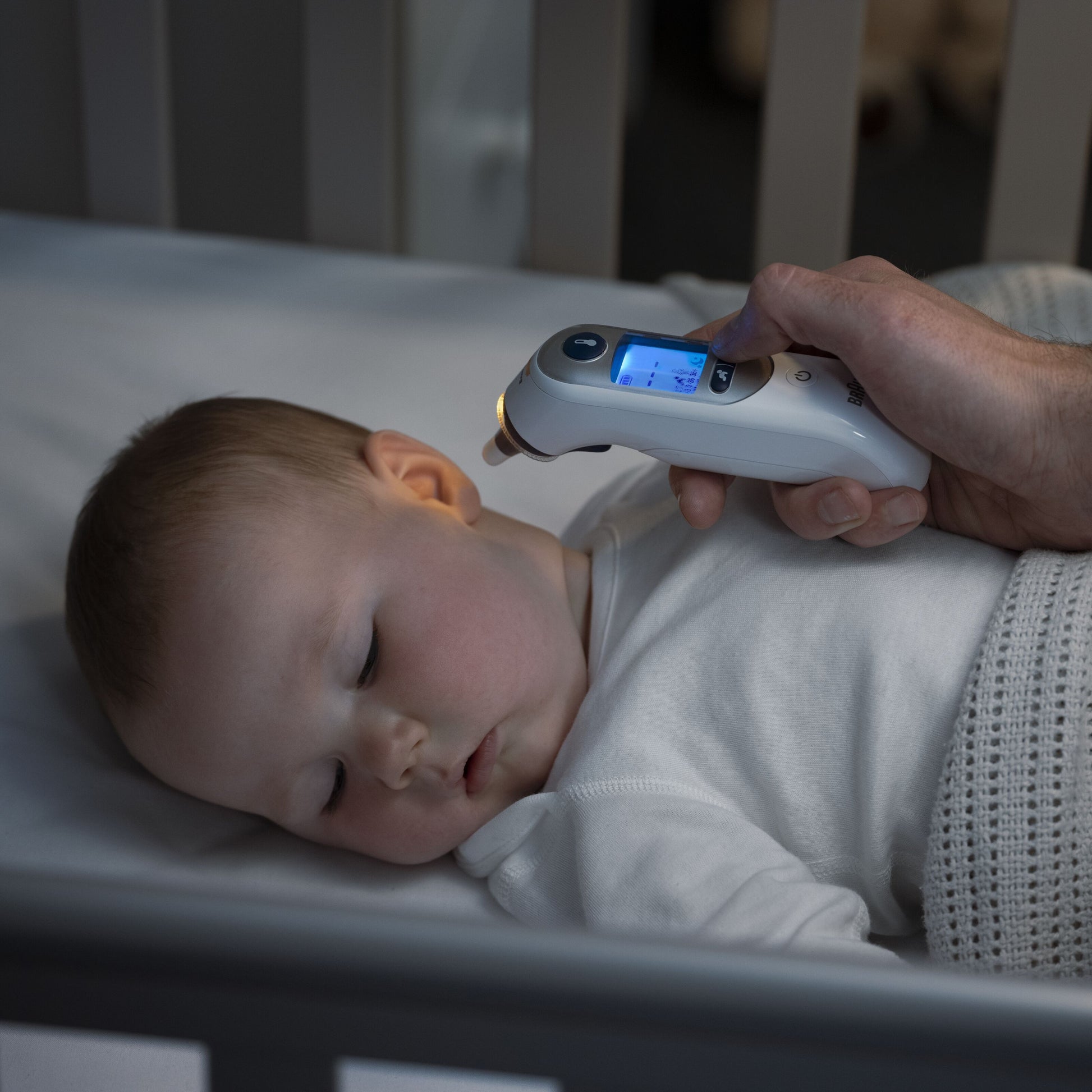 Braun Thermoscan 7+ Øretermometer bruges til at måle temperatur på baby i hvid sparkedragt og tæppe i tremmeseng.