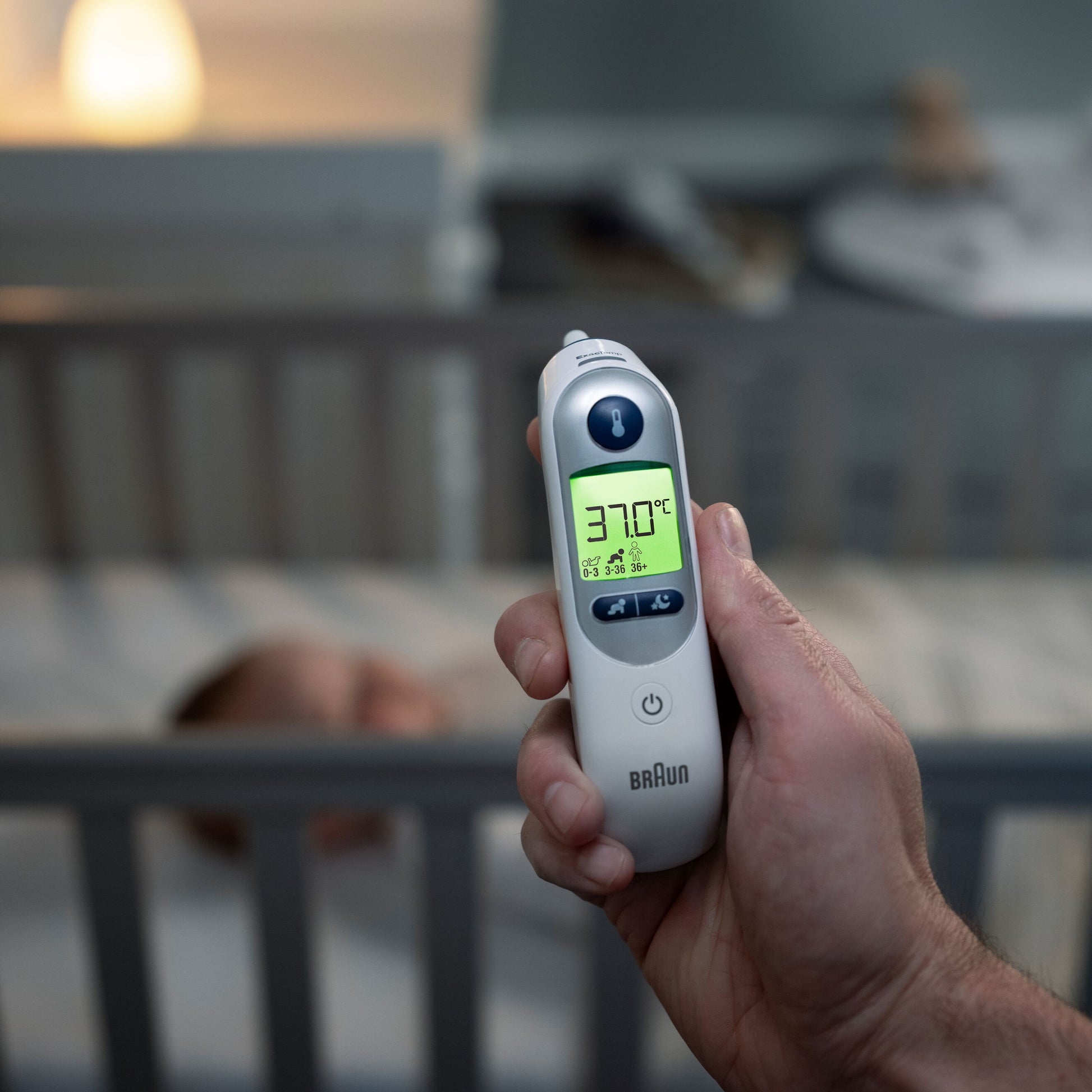 Braun Thermoscan 7+ øretermometer vises i hånden, display viser 37,0°C, sløret baby og tremmeseng i baggrunden.