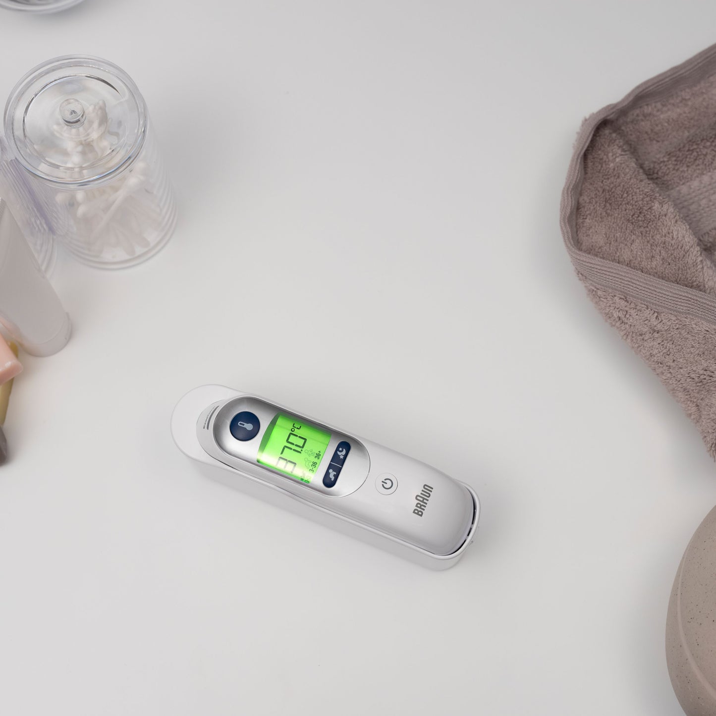 Braun Thermoscan 7+ øretermometer viser 37,0°C ved siden af vatpinde og sammenfoldet beige håndklæde.