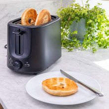 Brødrister Compact med to bagelhalvdele i, hvid tallerken med bagel og kniv ved siden af, grøn plante i baggrunden.