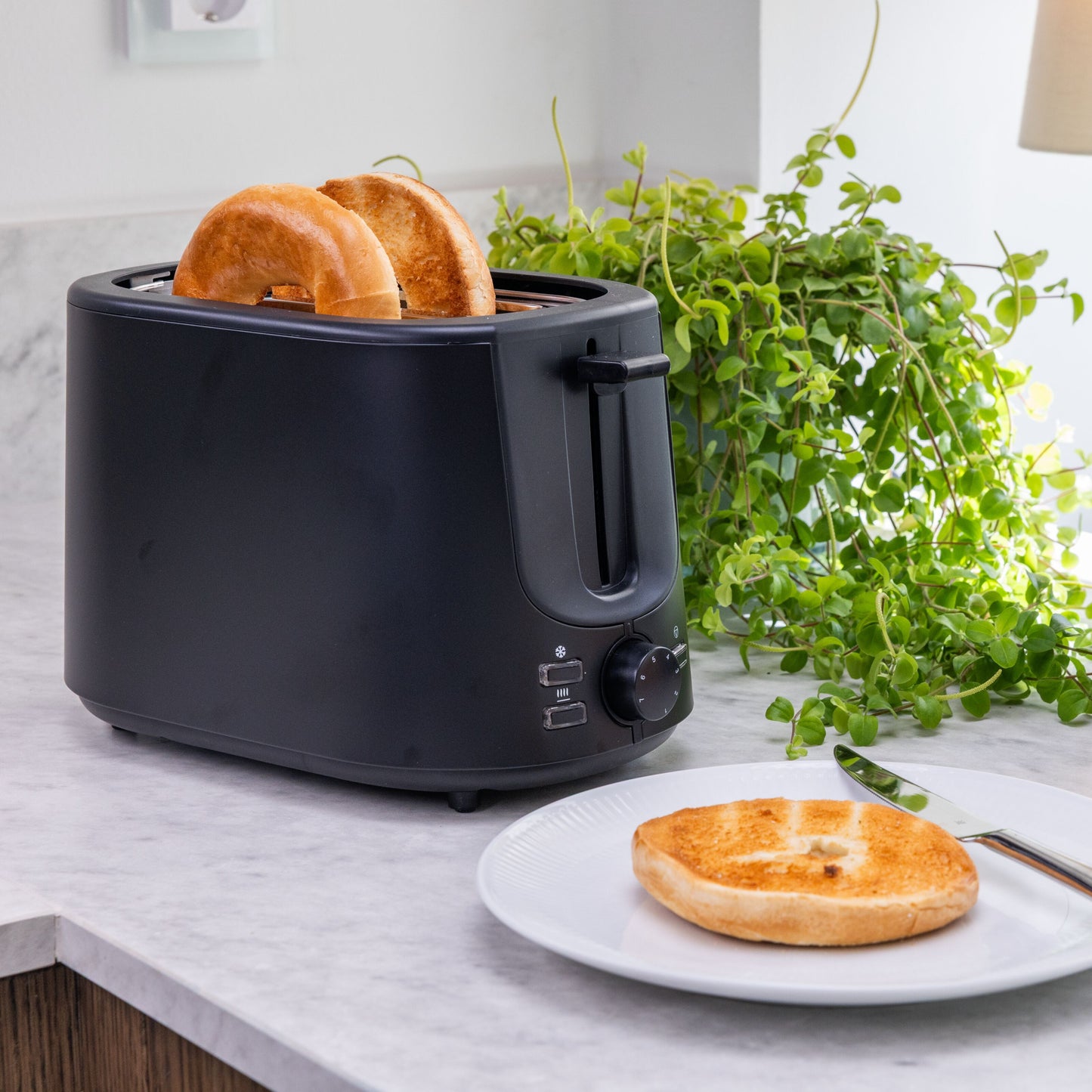 Brødrister Compact sort med 7 ristningsniveauer, toasting bagel, skive på hvid tallerken, grøn plante i baggrund.