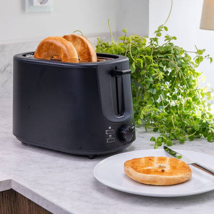 Brødrister Compact sort med 7 ristningsniveauer, toasting bagel, skive på hvid tallerken, grøn plante i baggrund.