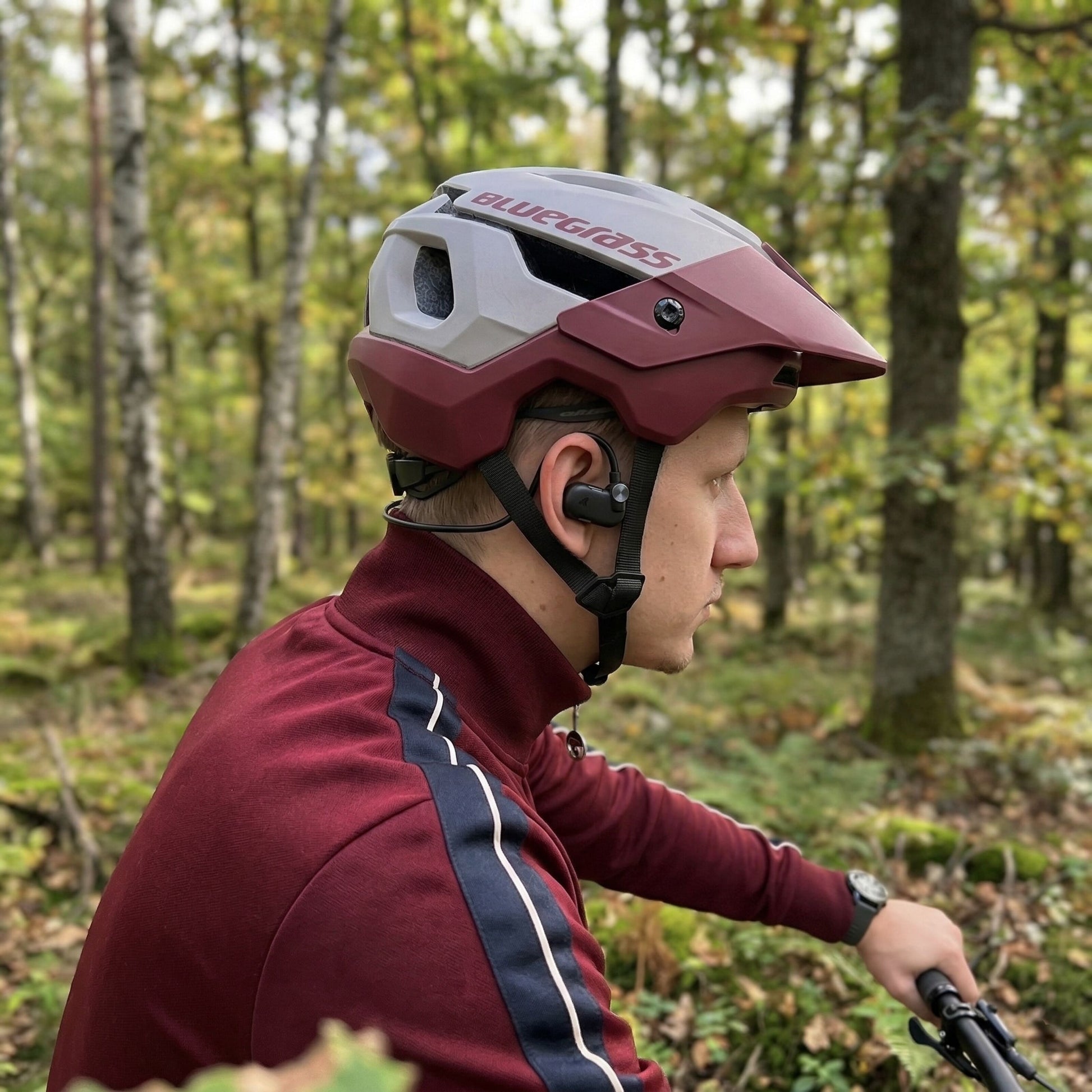 Nomadelic Solo 330 Sport open-ear-hovedtelefoner, rødbrun hjelm og jakke, vises udendørs.