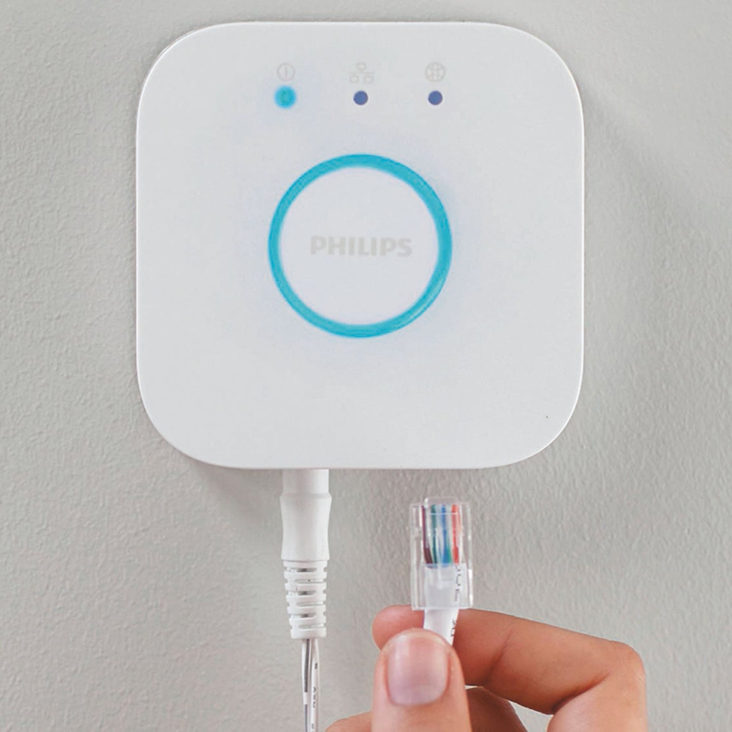 Philips Hue Bridge smart hub vægmonteret med blå lysring, statuslys og Ethernet-port, hånd holder netværkskabel.
