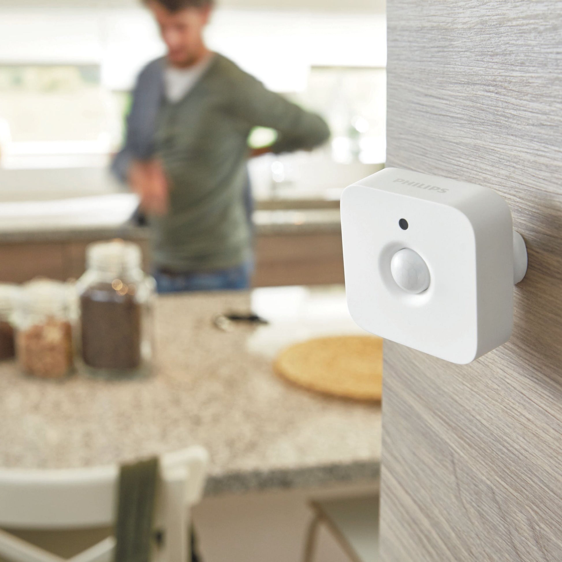Philips Hue bevægelsessensor til indendørs brug monteret på lyst træ i køkken, person sløret i baggrunden.