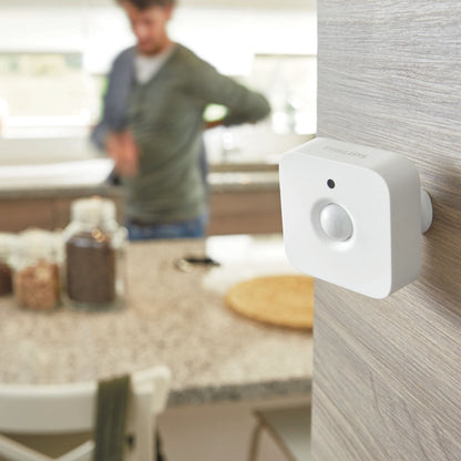 Philips Hue bevægelsessensor til indendørs brug monteret på lyst træ i køkken, person sløret i baggrunden.