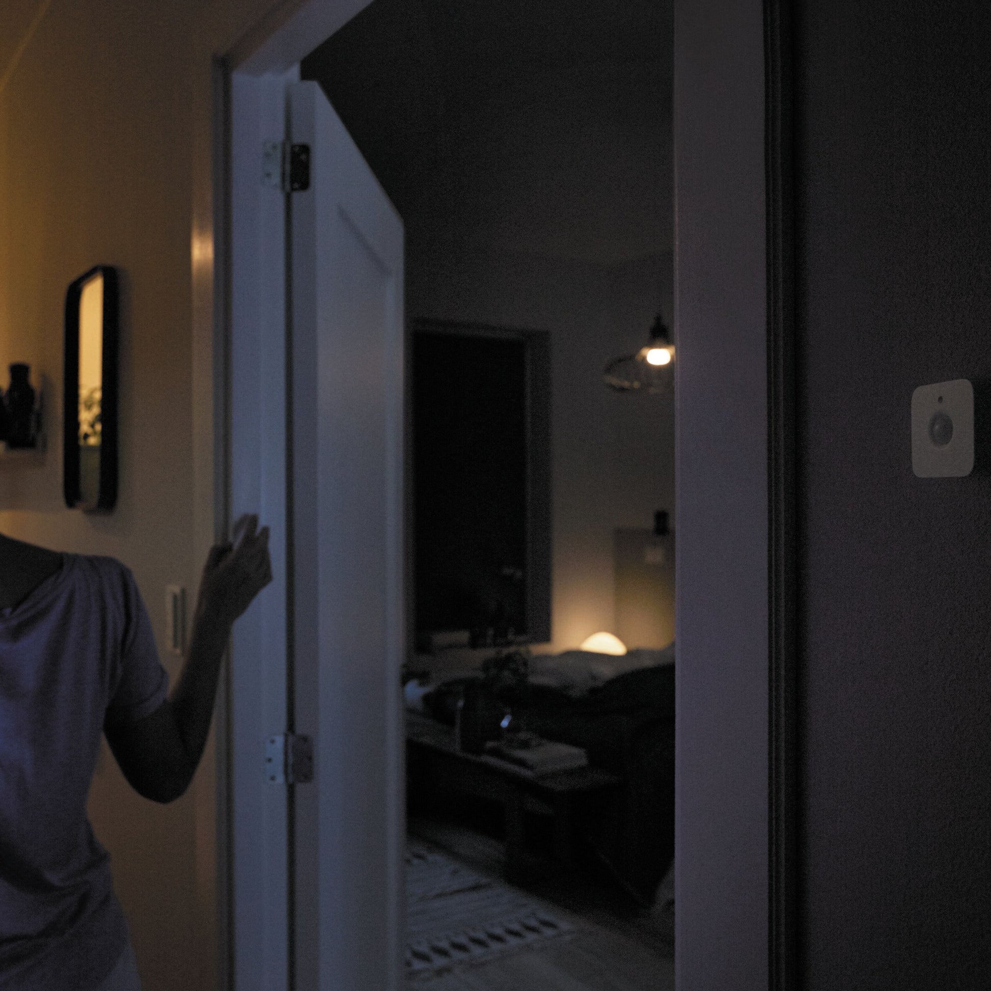 Philips Hue Motion sensor monteret på væg indendørs i svag belysning, person åbner dør iført lilla skjorte.
