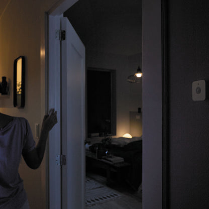 Philips Hue Motion sensor monteret på væg indendørs i svag belysning, person åbner dør iført lilla skjorte.