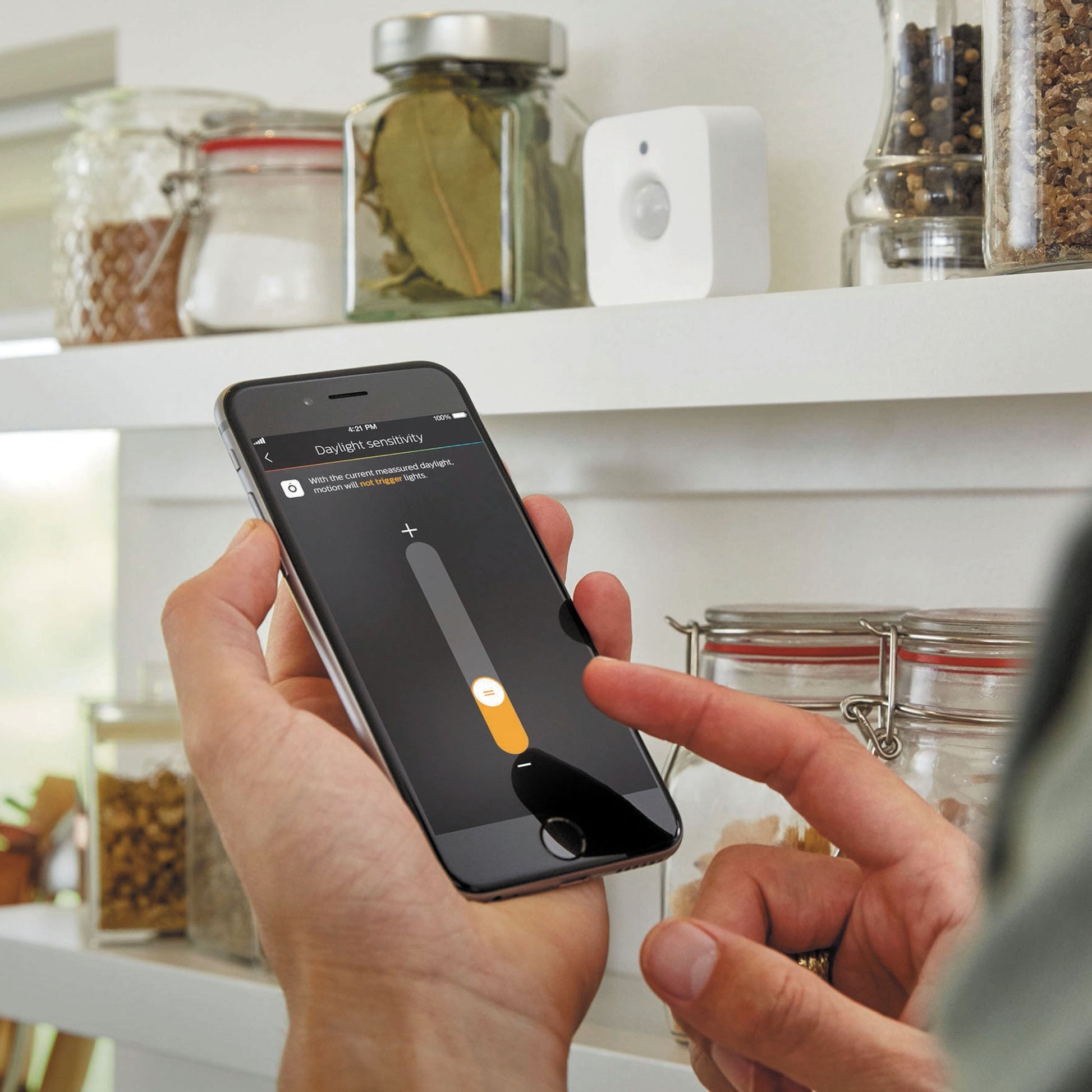 Philips Hue Motion sensor indendørs til intelligent belysning, synlig på køkkenhylde nær glas og krukker.