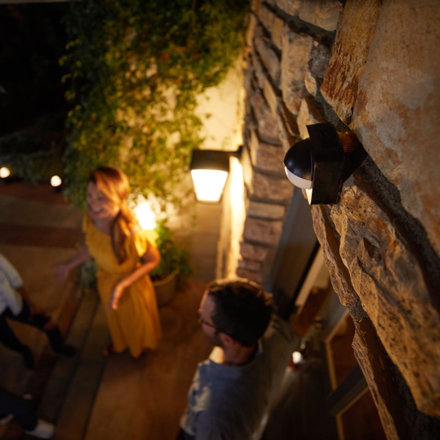 Philips Hue udendørs bevægelsessensor monteret på stenvæg ved lampe, personer synlige i baggrunden.
