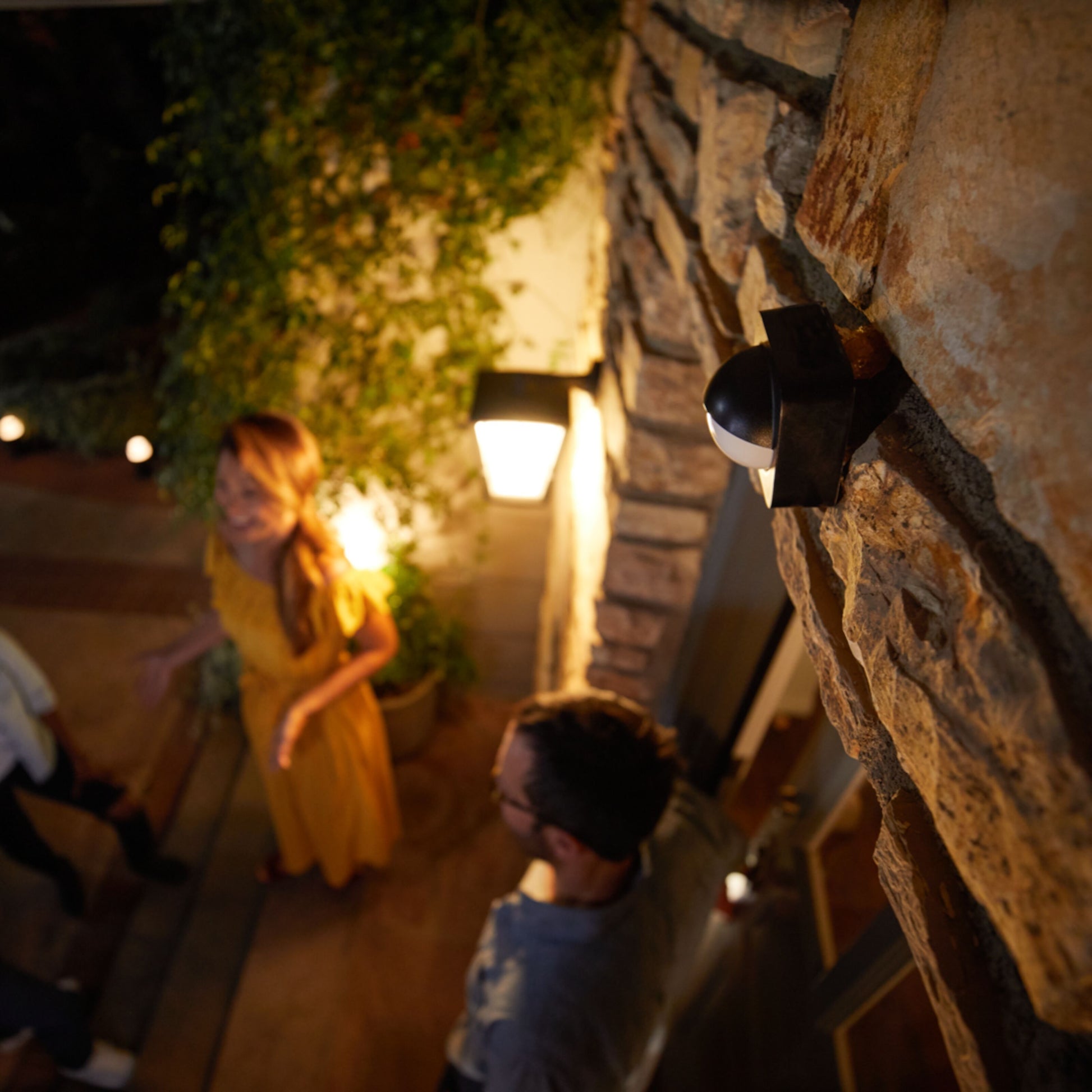 Philips Hue udendørs bevægelsessensor monteret på stenvæg ved lampe, personer synlige i baggrunden.