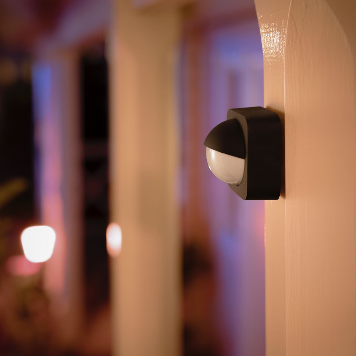 Philips Hue Bevægelsessensor sort til udendørs montering på beige væg ved dør, oplyst af udendørslys, aften.