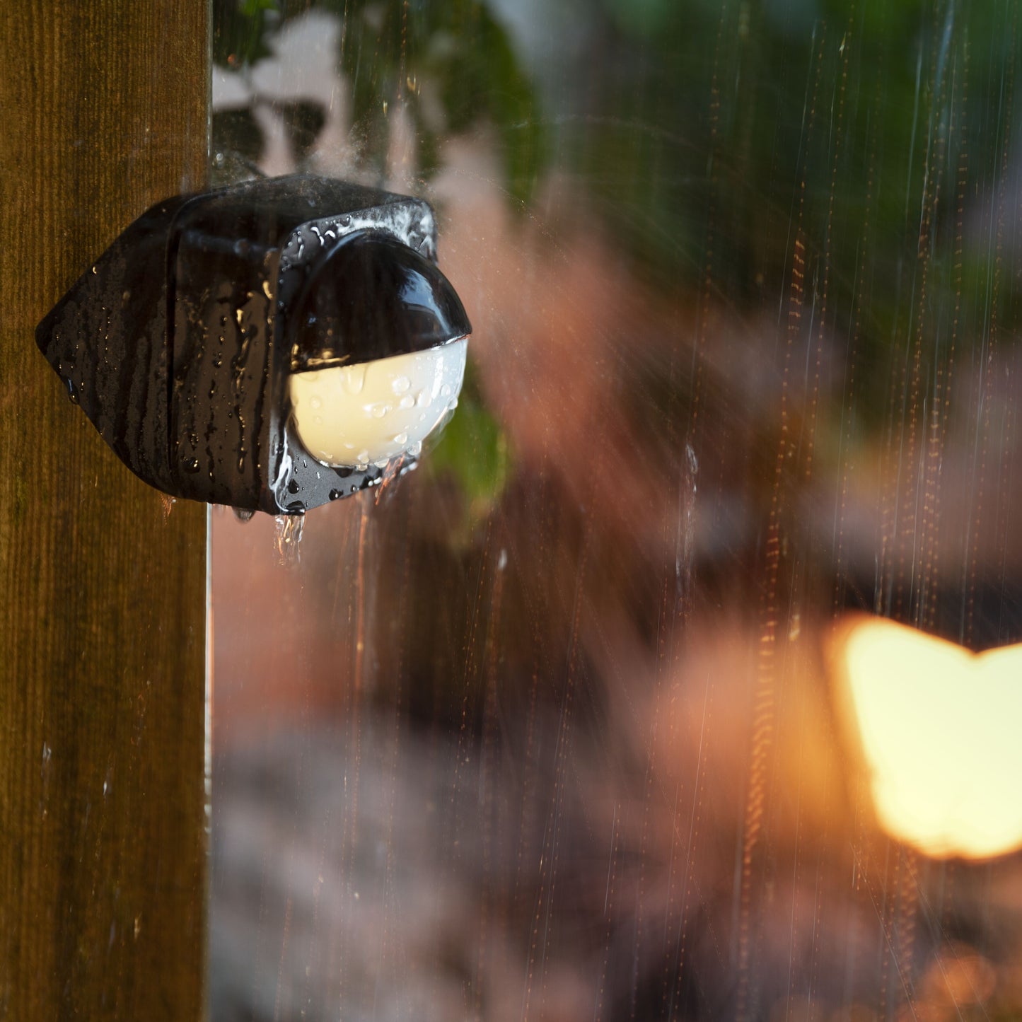 Philips Hue Bevægelsessensor til udendørs brug, våd med vanddråber, monteret på træstolpe i regn.