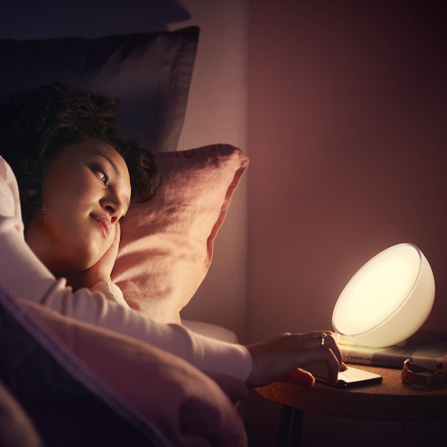 Philips Hue Go bærbar bordlampe lyser på natbord ved seng, synlig pink pude og Bluetooth-styring i brug.