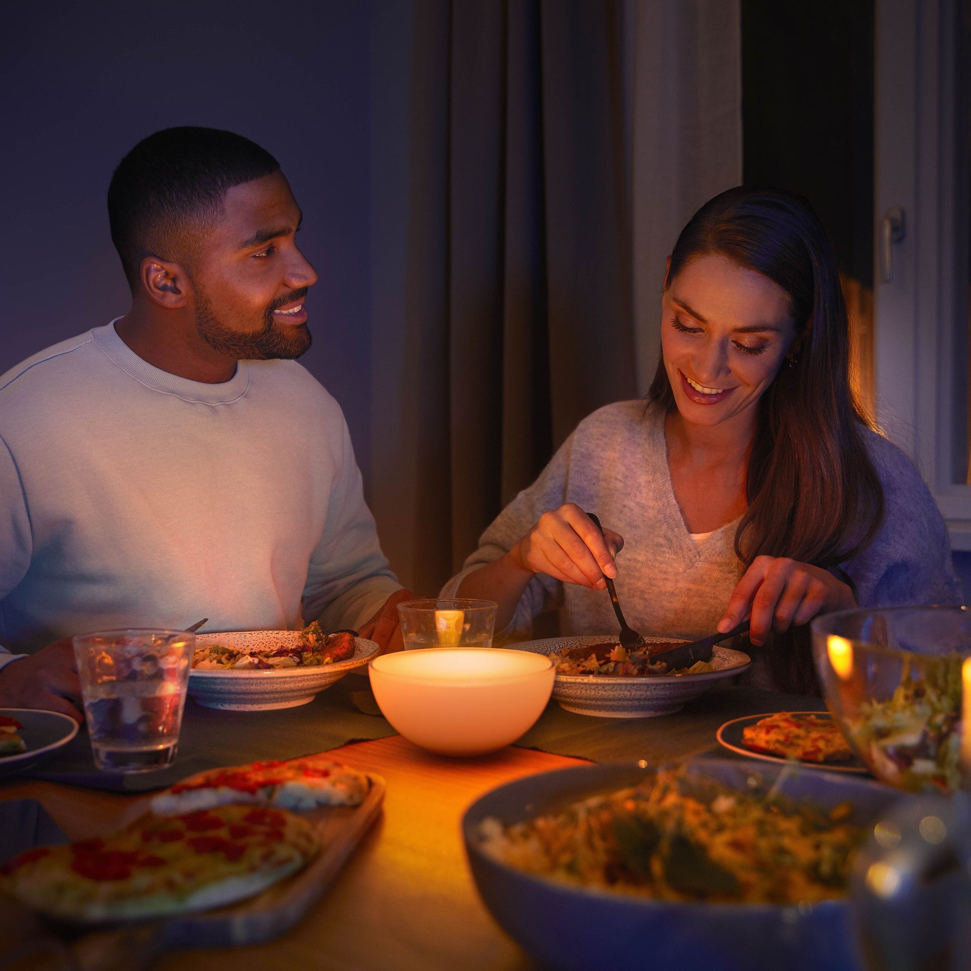 Philips Hue Go bærbar bordlampe med Bluetooth ses tændt på spisebord mellem to personer, rund model, hvid.