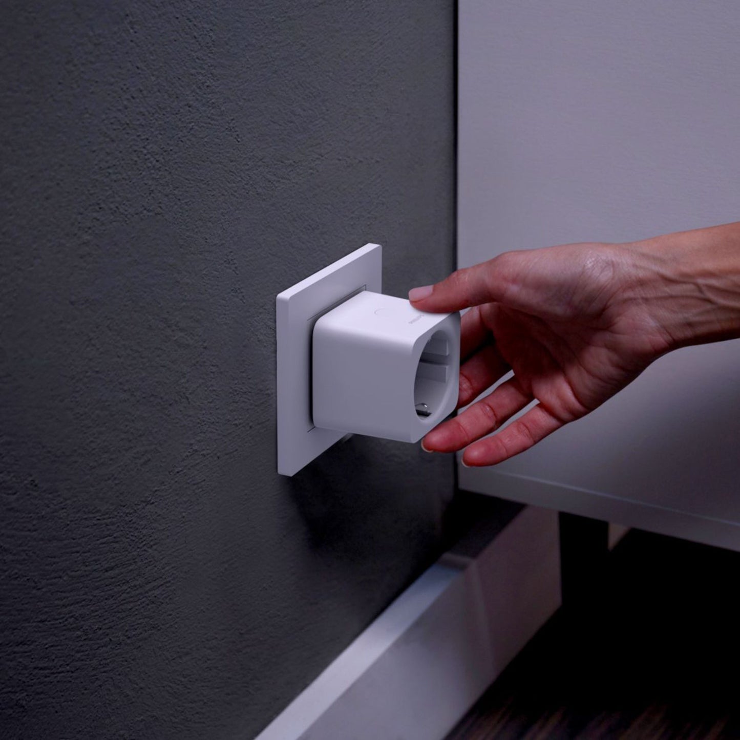 Philips Hue Smart Plug fjernbetjent strømafbryder sættes i stikkontakt på grå væg ved siden af hvidt møbel.