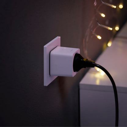 Philips Hue Smart Plug fjernbetjent strømafbryder sort i brug ved hvidt møbel, varm lyssætning på mørk væg.